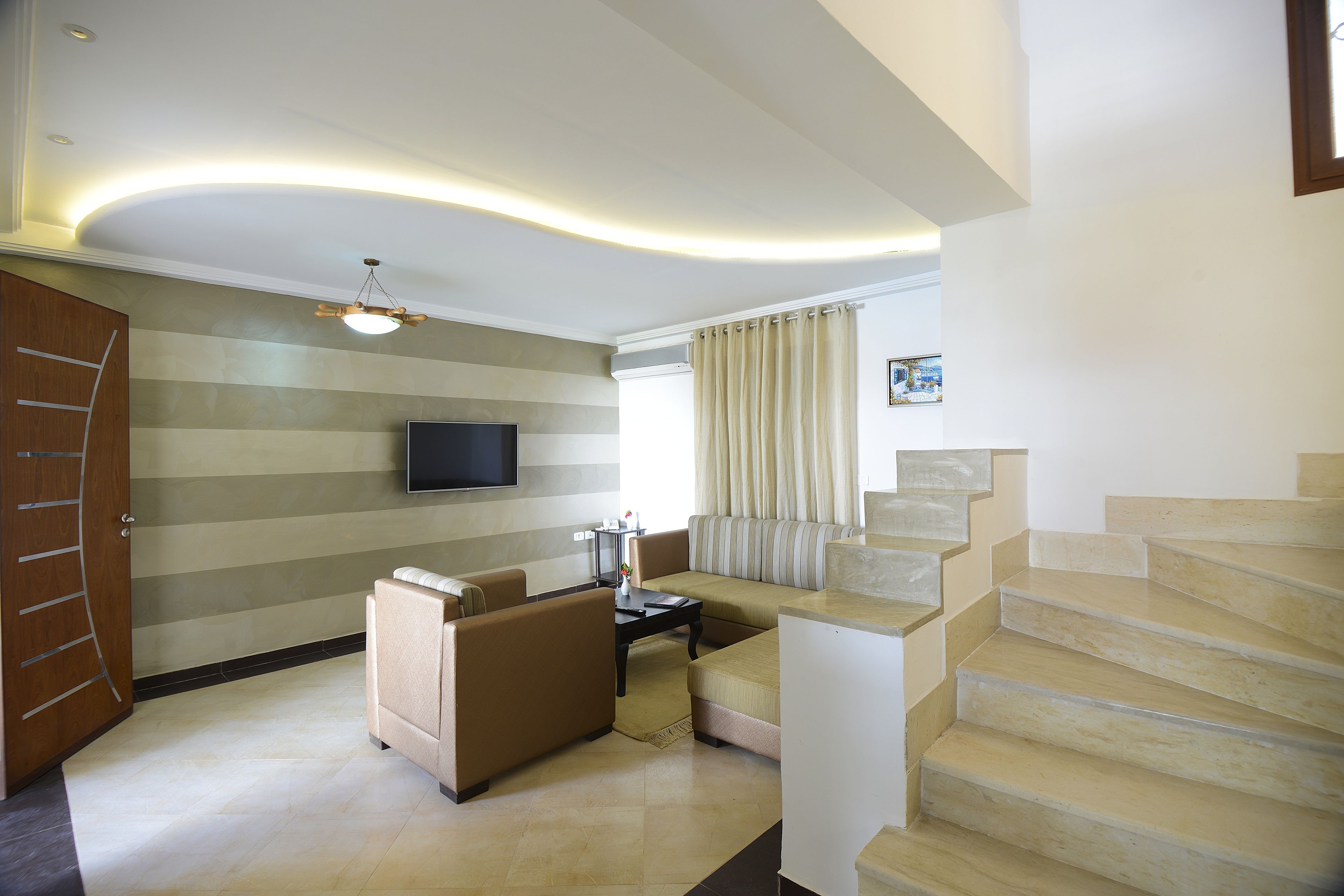 A modern living room with a sofa, a coffee table, and a flat-screen TV mounted on the wall. There is a staircase with beige steps leading to another level.