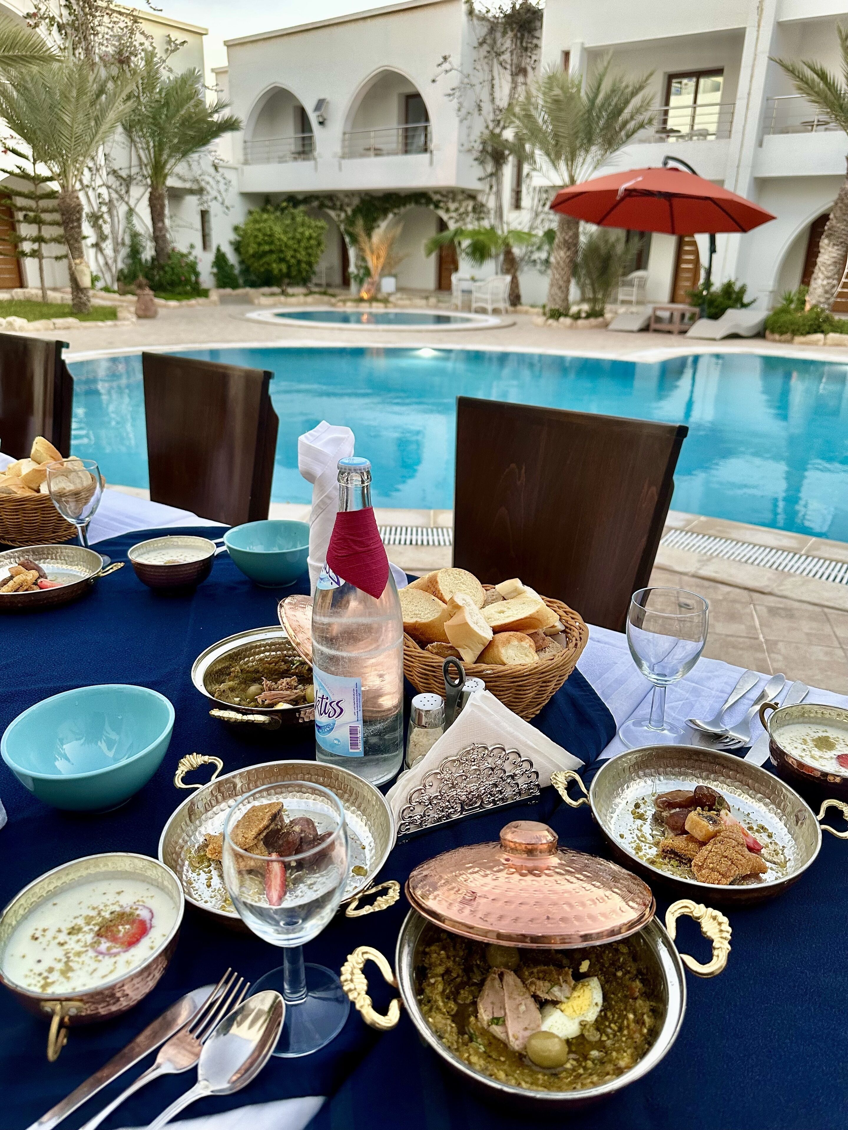 A dining table set with various dishes, a bottle of water, and a red umbrella near a poolside.