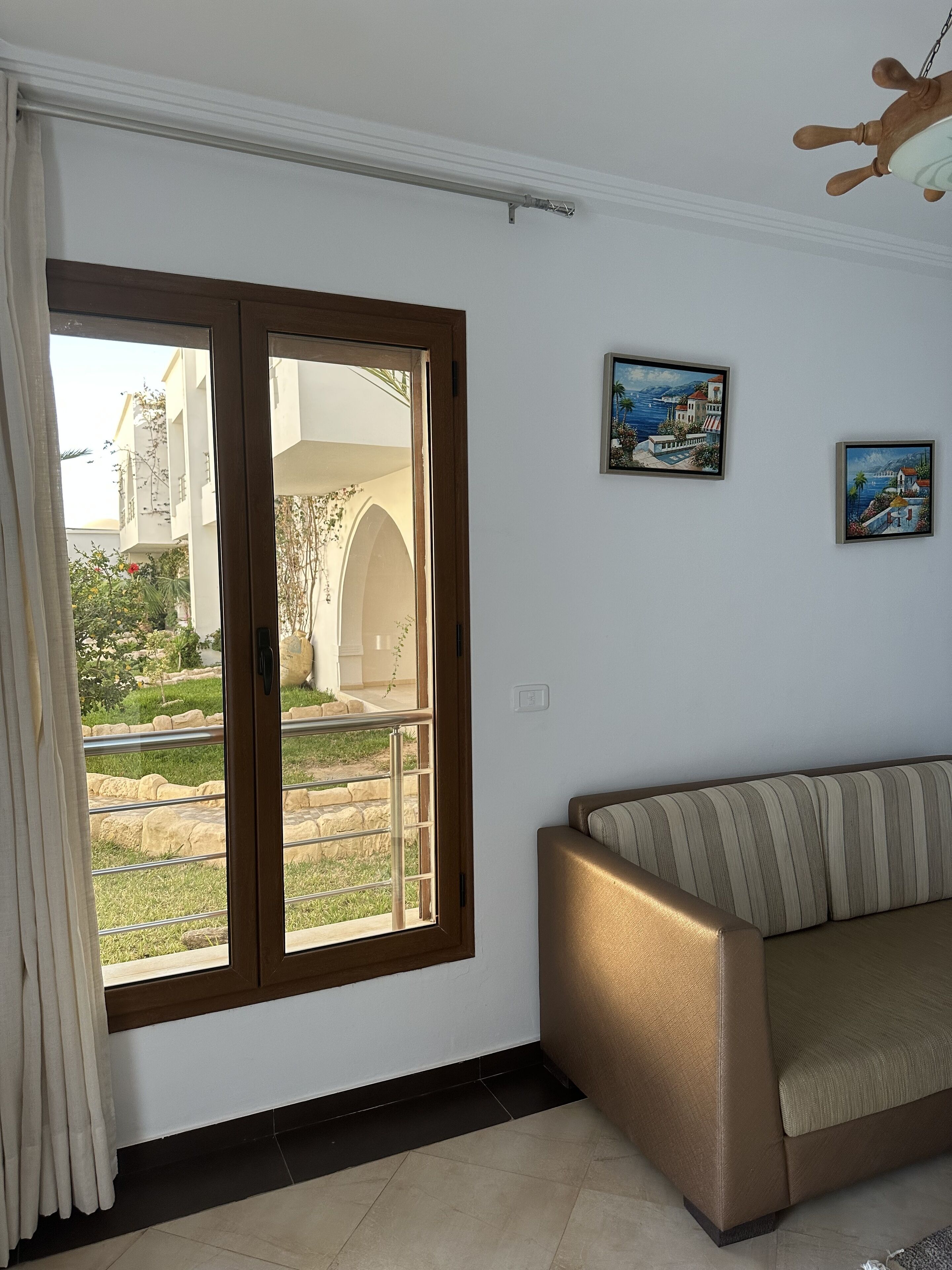 A room with a sofa, a large window with a view of a building and greenery, and two framed pictures on the wall.