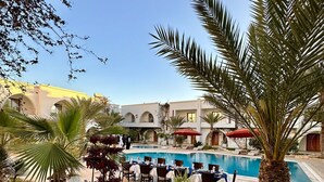A poolside area with outdoor seating and a pool, surrounded by palm trees and a building with arches. - Palm Djerba Suites (Mezraia)