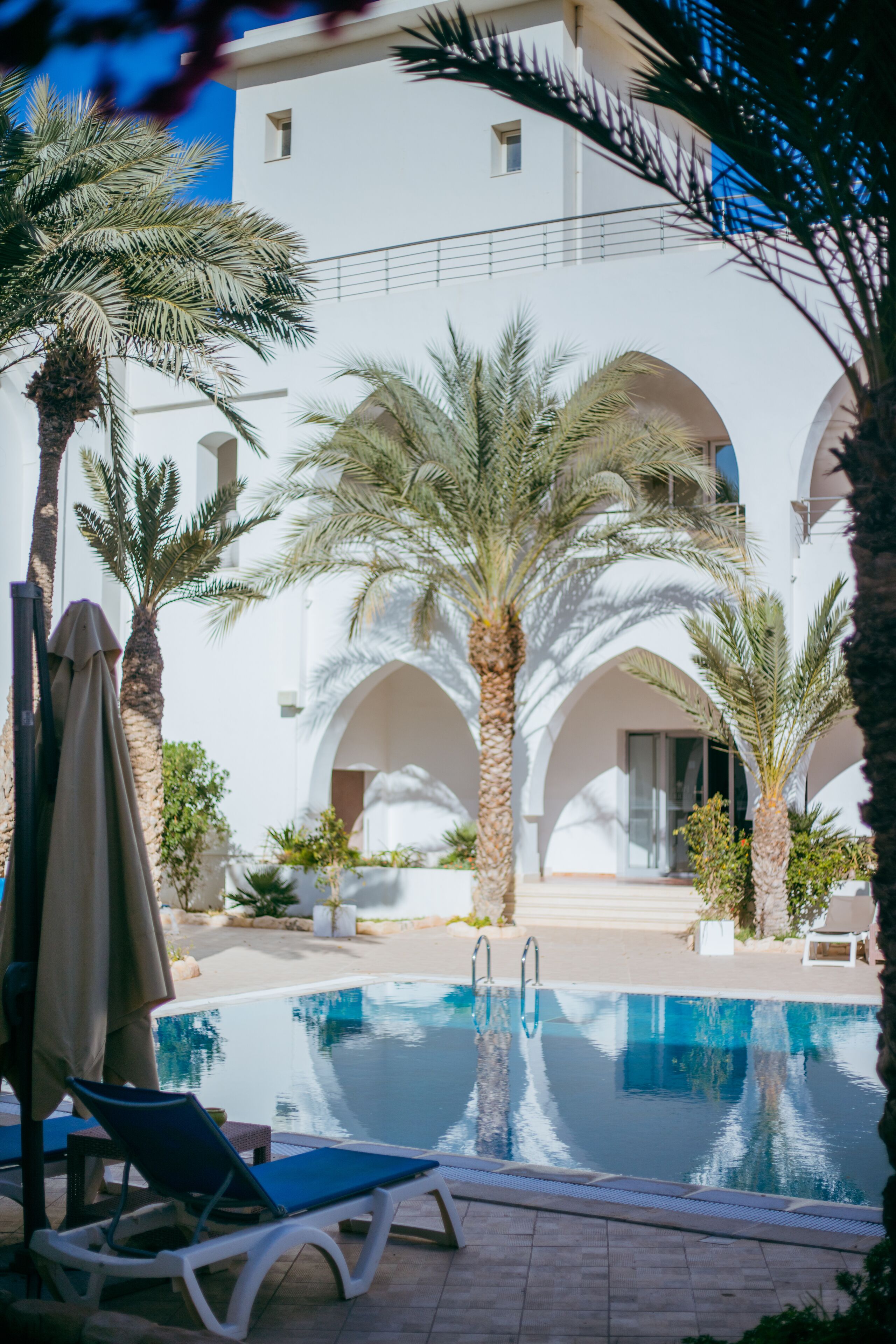 A pool area with lounge chairs, palm trees, and a white building with arches.