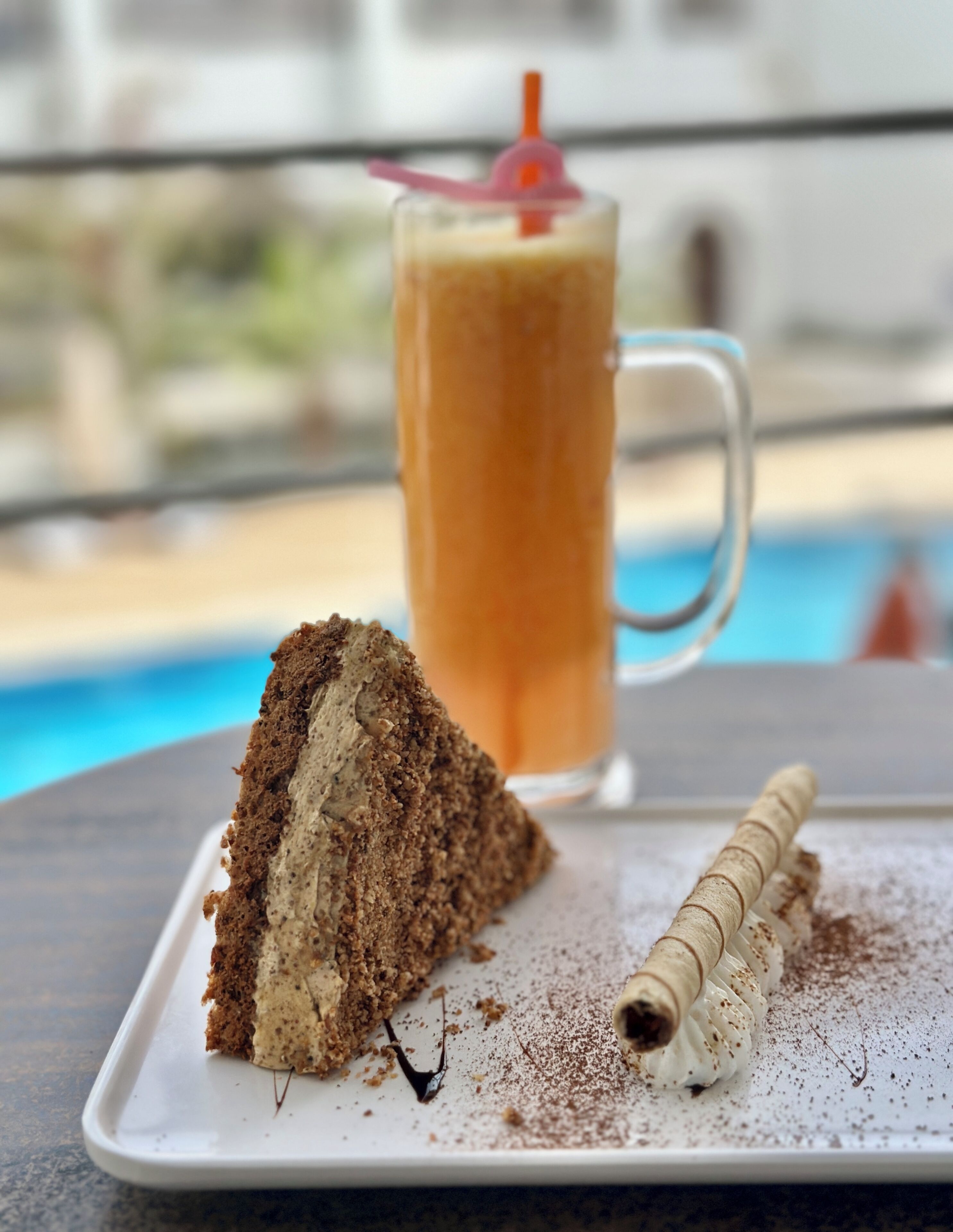Una fetta di torta con ripieno cremoso e una spolverata di cacao amaro, un bastoncino wafer arrotolato e un bicchiere alto di caffè freddo con una cannuccia su un piatto bianco.