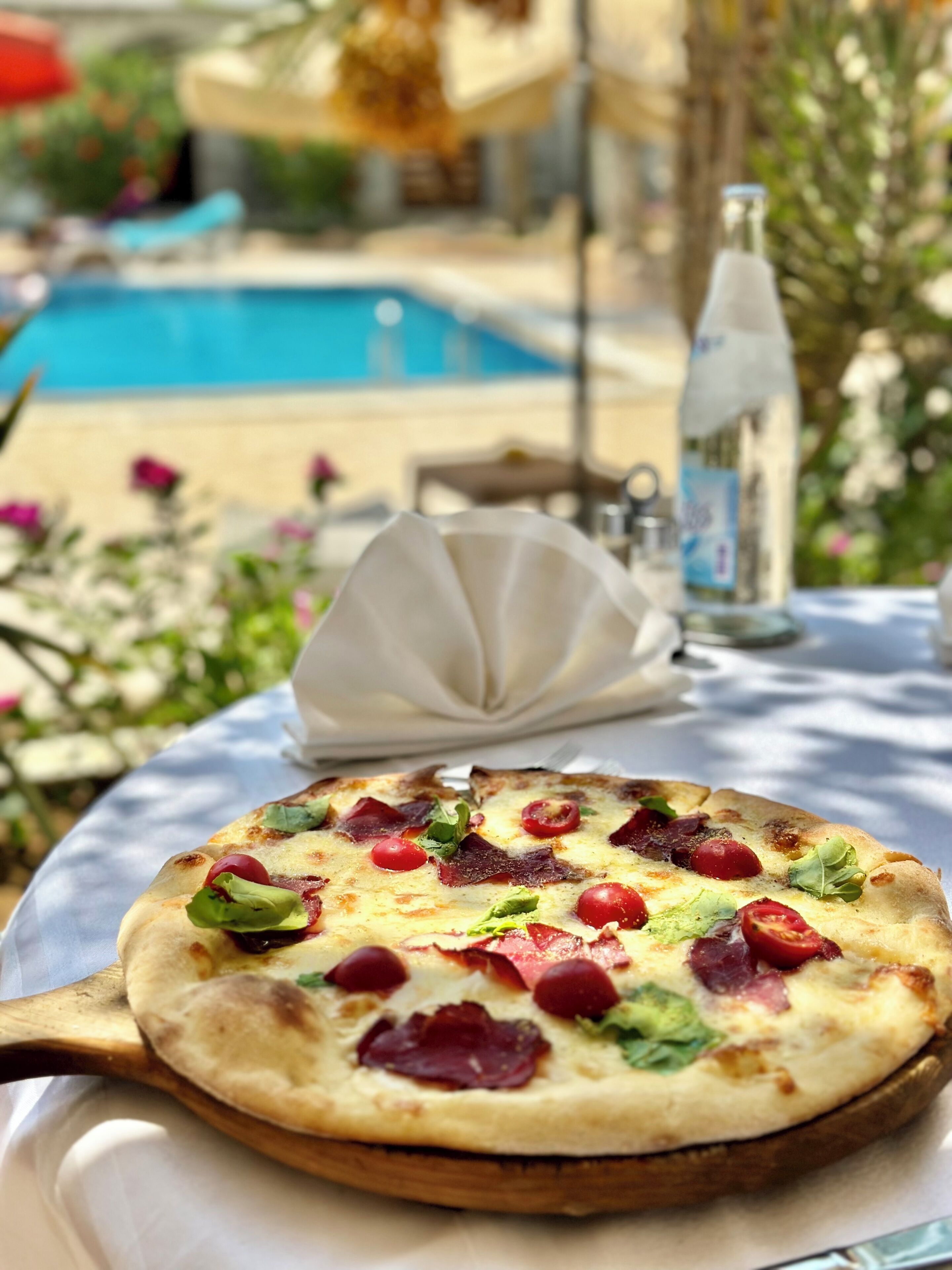 Pizza auf einem Tisch, im Hintergrund ein Schwimmbad und eine Flasche.