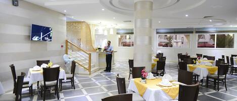 A hotel dining area with checkered flooring, multiple tables with yellow tablecloths, and dark wooden chairs.
