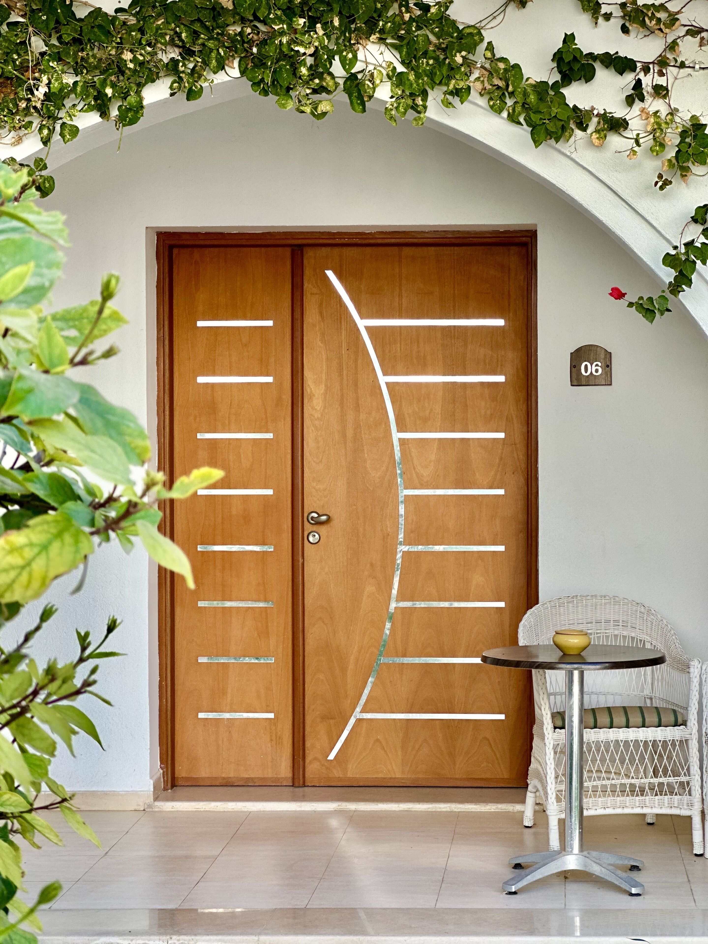 A wooden double door with a unique curved design, a small round table with a yellow object, and a white wicker chair.