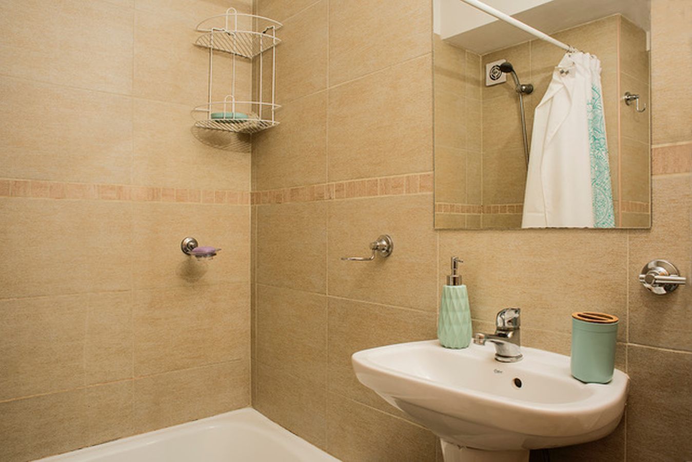 A bathroom with beige tiles, a white sink, a mirror, a shower, and a hanging towel rack.