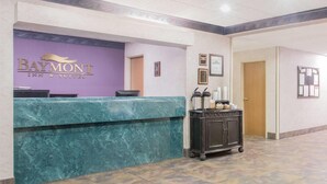 A reception area with a marble counter, a Baymont Inn & Suites sign, framed certificates, and a cart with amenities. - Baymont Inn and Suites (Corbin)