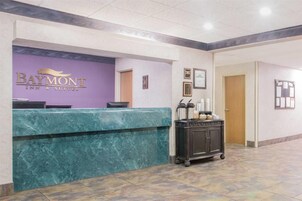 Zona de recepción con mostrador de mármol, cartel de Baymont Inn & Suites, certificados enmarcados y un carrito con servicios.
