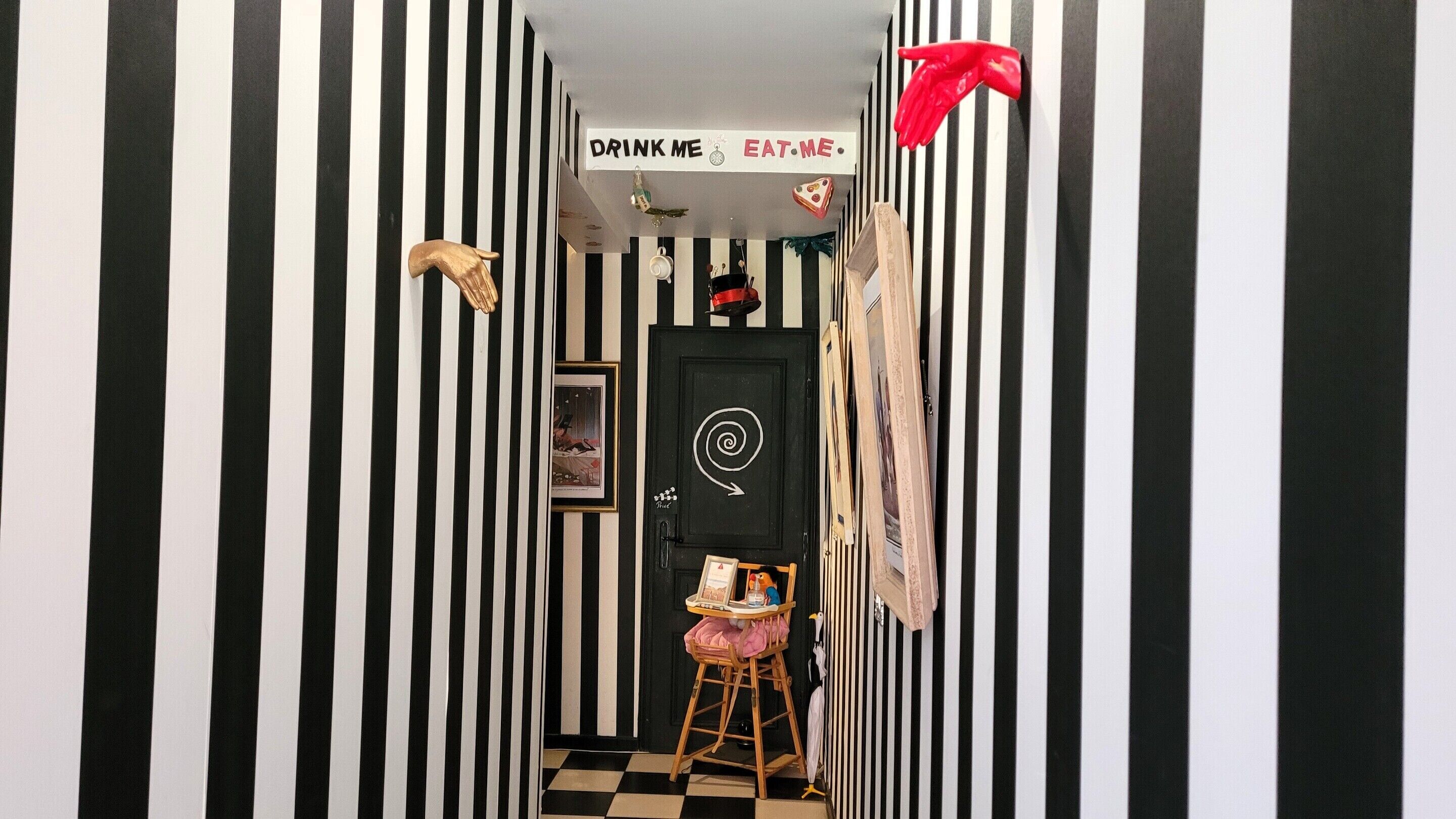 A narrow hallway with black and white striped walls, a sign above the door that reads 'DRINK ME EAT ME', and a small chair with a pink cushion.