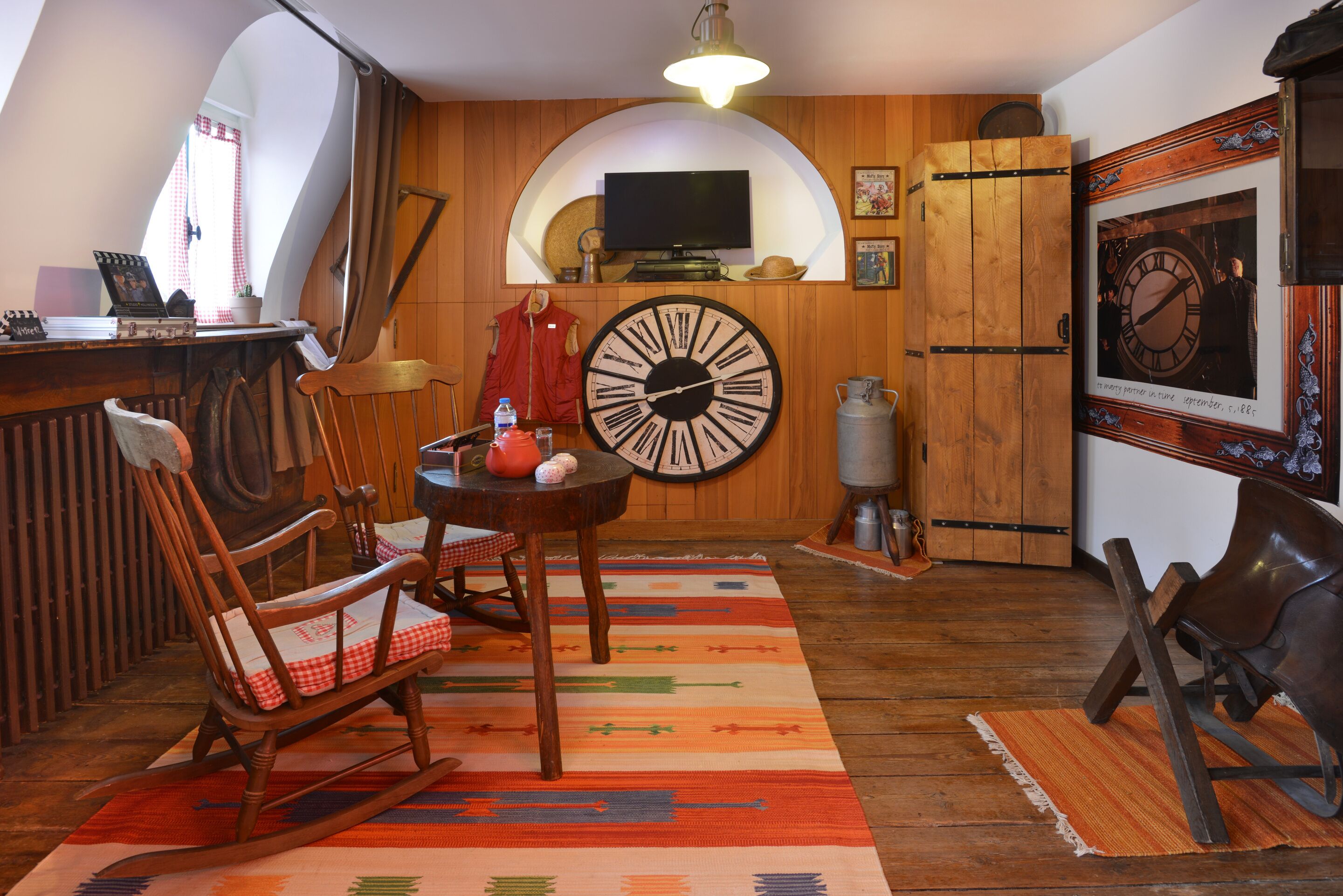 A cozy room with a wooden floor, a rocking chair, a table with a red and white checkered tablecloth, a large wall clock, and a television on a wooden shelf.