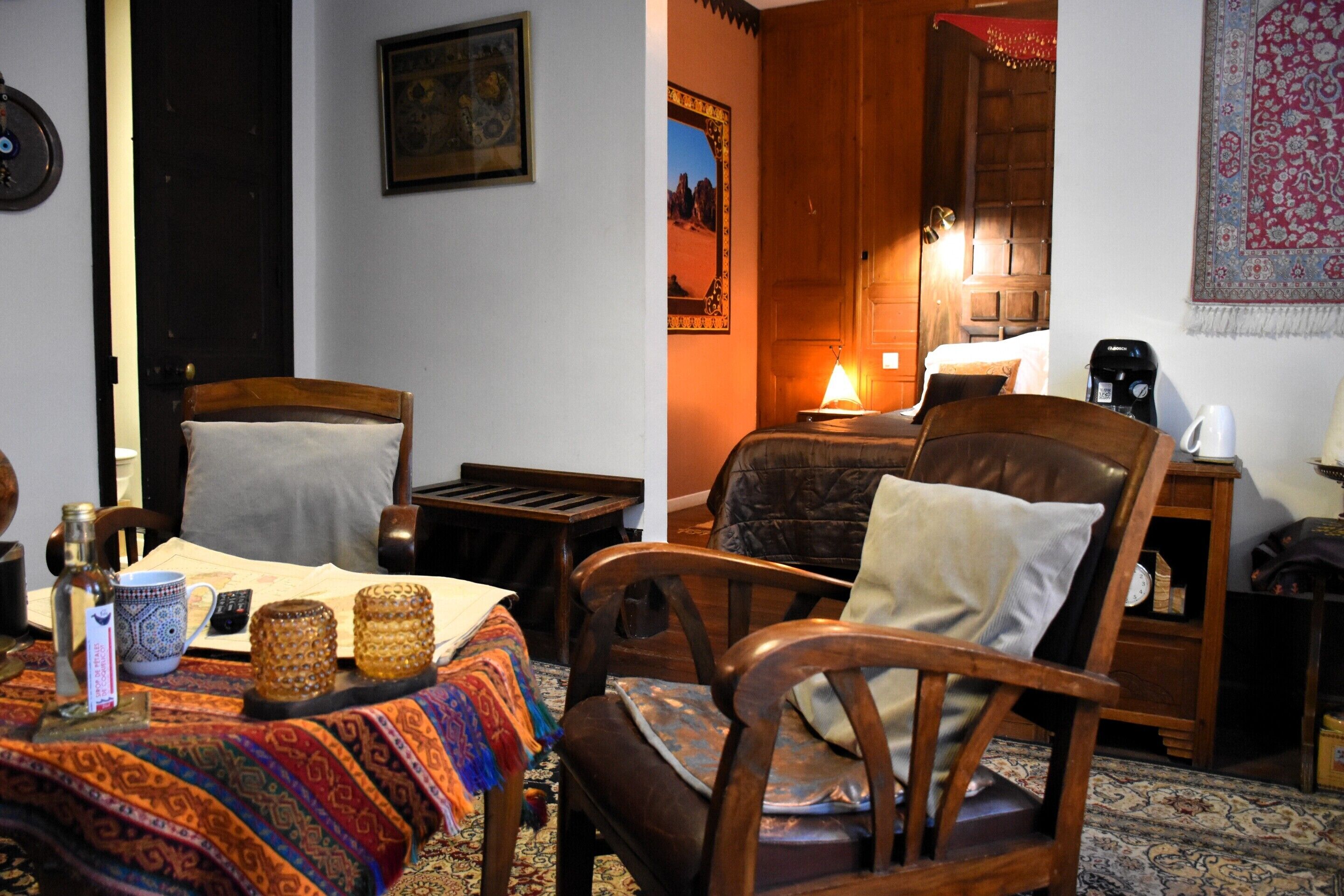 Una habitación acogedora con una cama, dos sillas de madera, una mesa con mantel colorido, una lámpara y un cuadro colgado en la pared.
