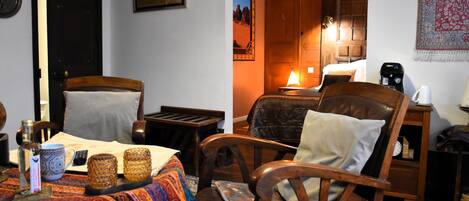 Una habitación acogedora con una cama, dos sillas de madera, una mesa con mantel colorido, una lámpara y un cuadro colgado en la pared.