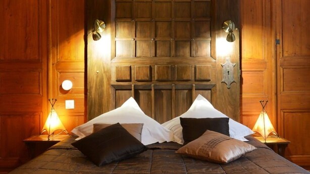 A bed with a dark brown quilt and multiple pillows, flanked by wooden walls and two wall-mounted lamps.