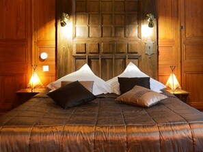 A bed with a dark brown quilt and multiple pillows, flanked by wooden walls and two wall-mounted lamps.