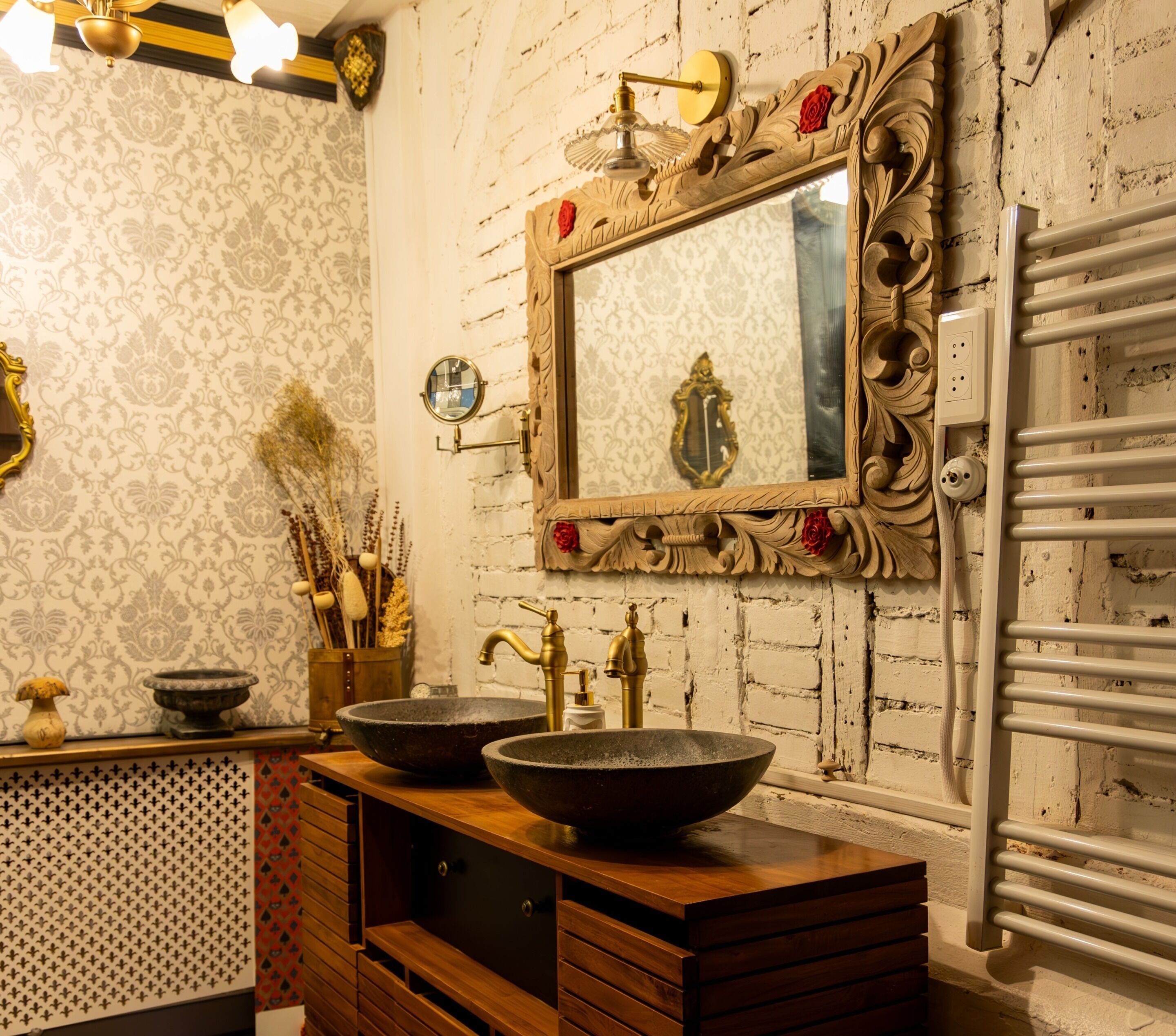 Un baño de estilo rústico, con un tocador de madera y dos lavamanos de piedra, un espejo con detalles ornamentales y un radiador blanco.