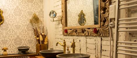 A bathroom with a rustic design, featuring a wooden vanity with two large stone sinks, a mirror with ornate carvings, and a white radiator.