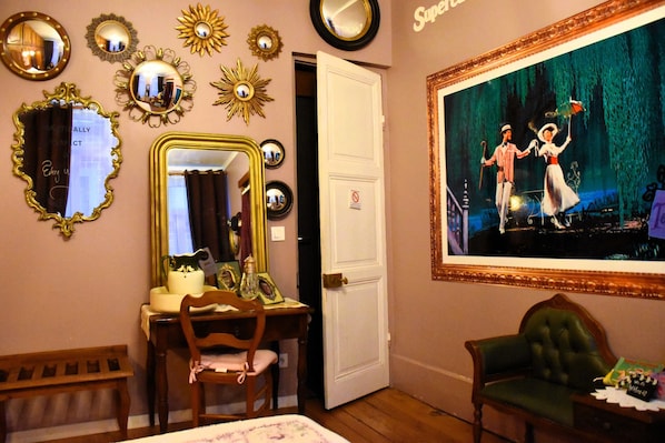 A room with a desk, chair, and a large mirror. There is a door and a framed picture on the wall.