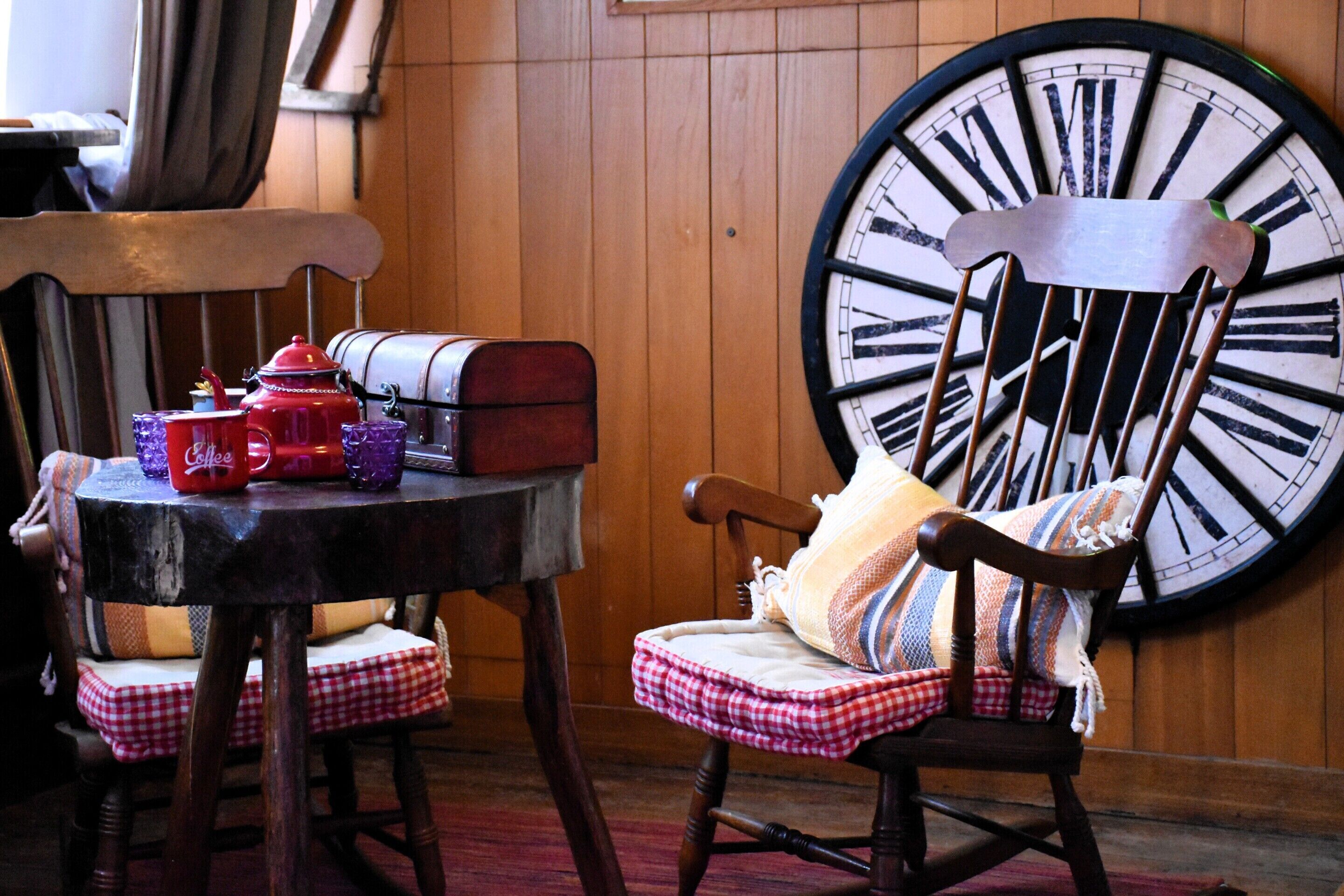 A wooden interior with a large clock, a wooden chair with a checkered cushion, and a small table with a red mug and a suitcase.