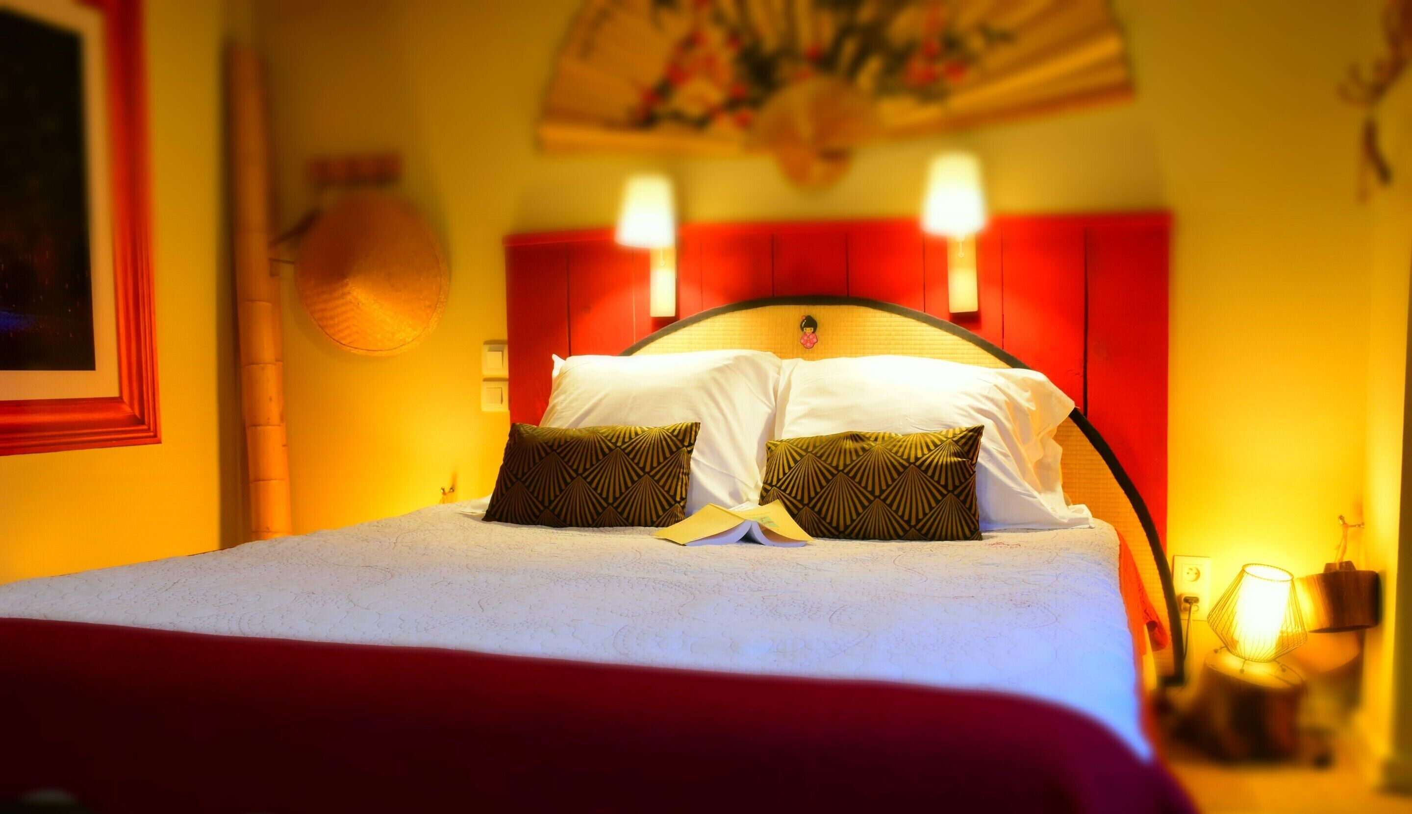 A neatly made bed with white linens and two patterned pillows, a red headboard, and two wall-mounted lamps.