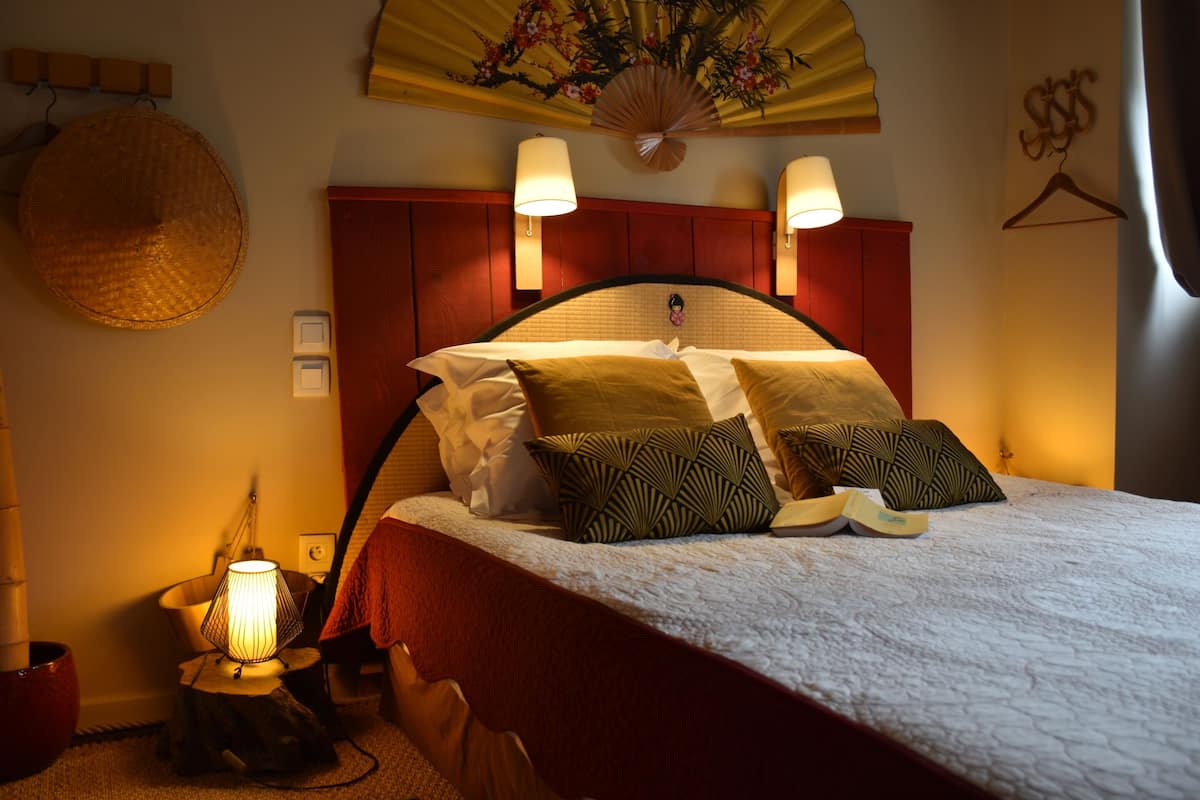 Una cama bien hecha con un cabecero rojo, dos lámparas de pared y un abanico decorativo en el techo.