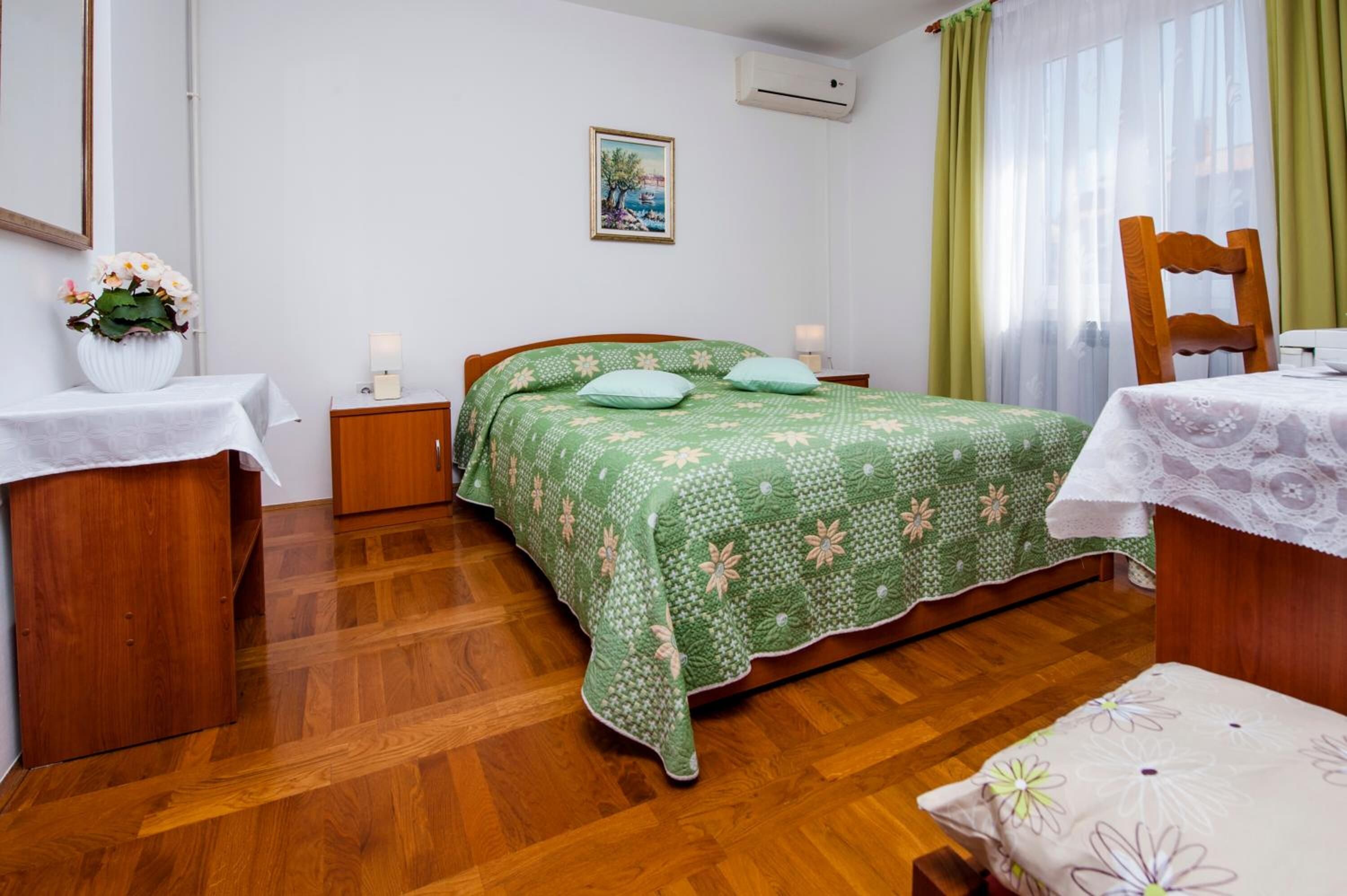 A bedroom with a wooden floor, a bed with a green floral bedspread, a nightstand with a lamp, a chair, and a table with a lace tablecloth.
