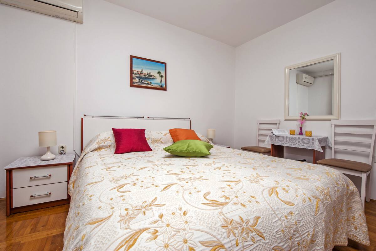 Un dormitorio ordenado con cama, mesitas de noche, tocador y una pequeña mesa con objetos decorativos.
