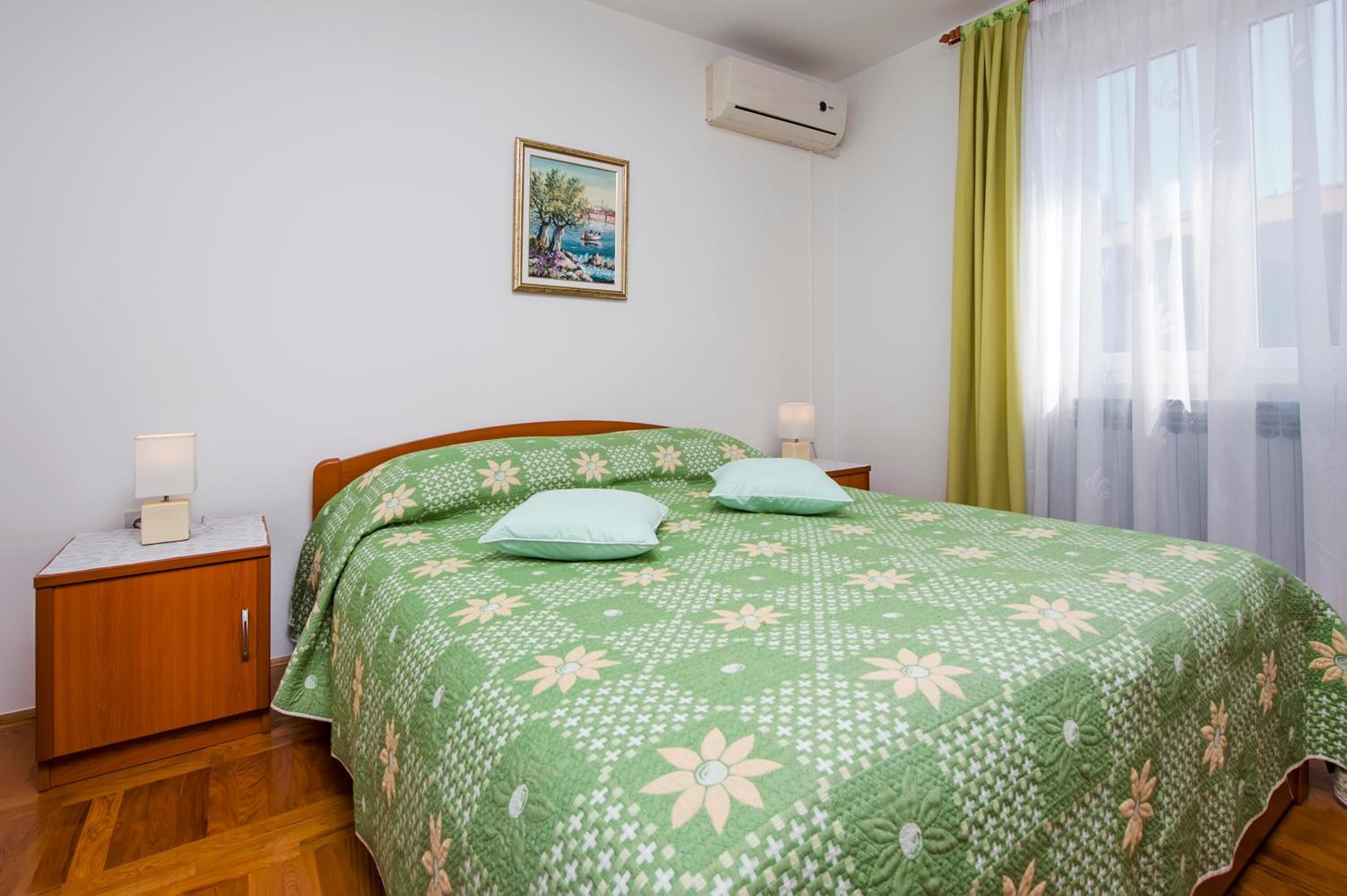 A bedroom with a green floral bedspread, two bedside lamps, a nightstand, a framed picture on the wall, and a window with sheer curtains.