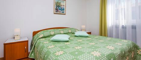 A bedroom with a green floral bedspread, two bedside lamps, a nightstand, a framed picture on the wall, and a window with sheer curtains.