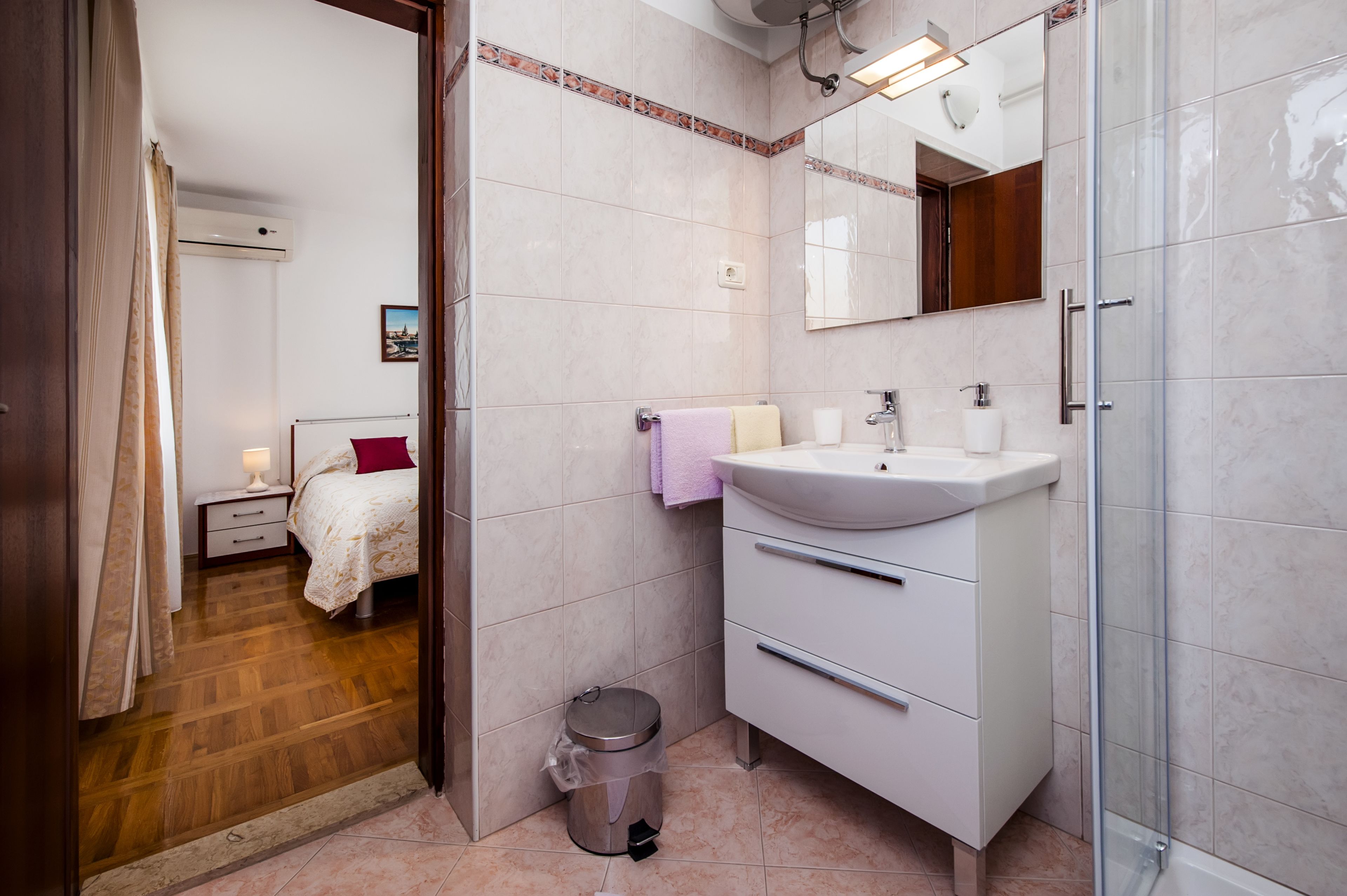Une salle de bain moderne équipée d’une douche, d’un lavabo et de toilettes. On y trouve une petite poubelle et une table de chevet avec une lampe.