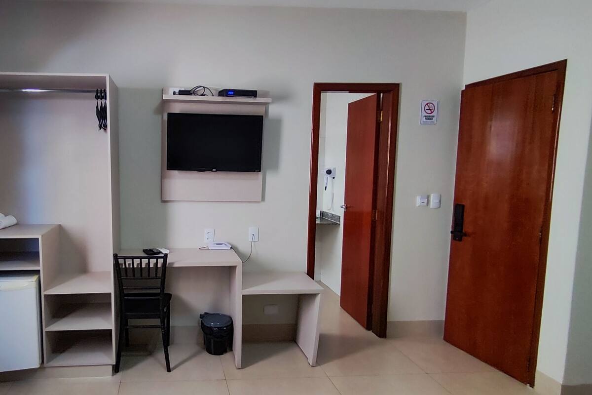 A compact room with a desk, a chair, a small refrigerator, a TV mounted on the wall, and a wardrobe.