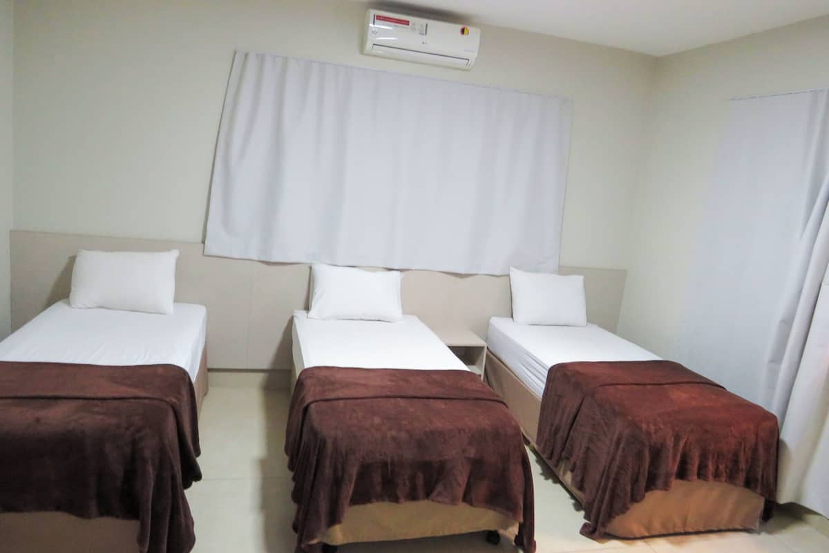 A hotel room with three single beds, each with a brown blanket, white pillows, and a white headboard. There is a white air conditioning unit on the wall above the beds.