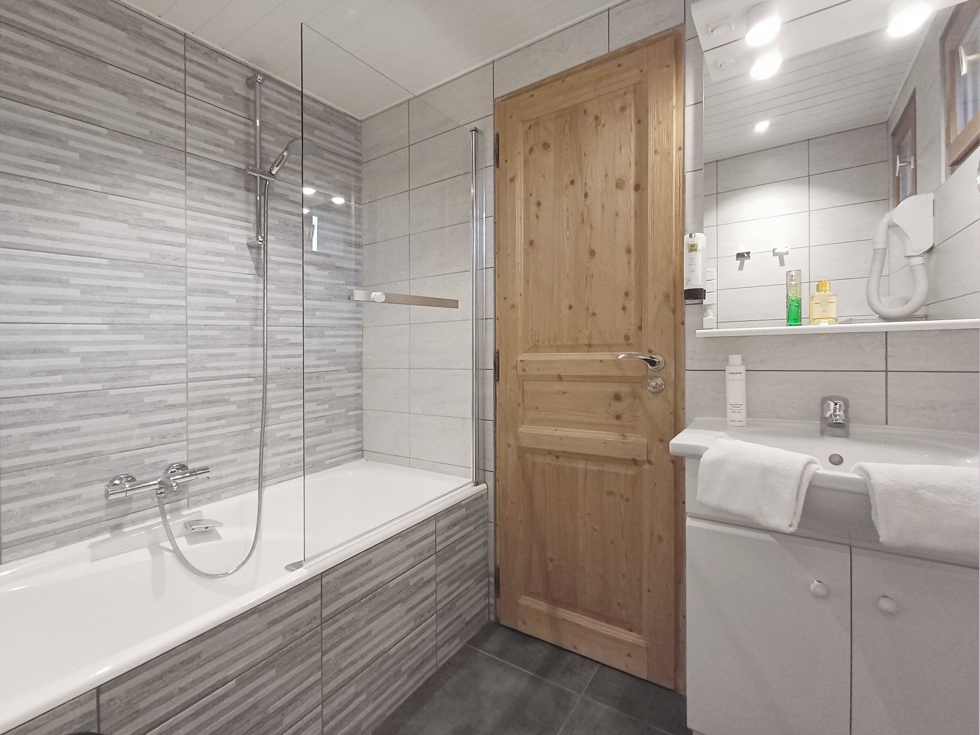 A modern bathroom with a glass-enclosed shower, a wooden door, a white sink, and a tiled shower area.