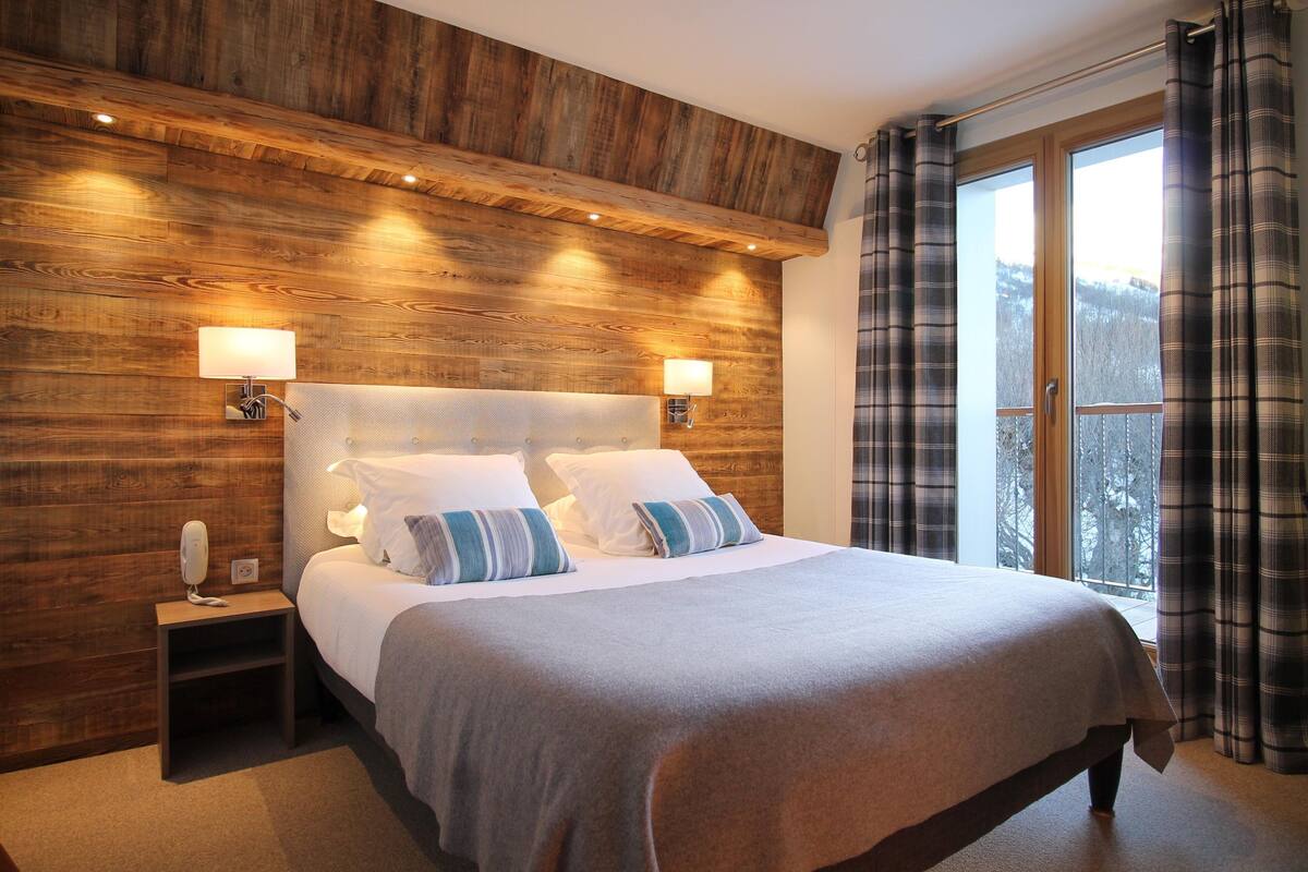 A bedroom with a wooden headboard, two bedside lamps, a bed with white and blue striped pillows, and a view of trees through a window with plaid curtains.