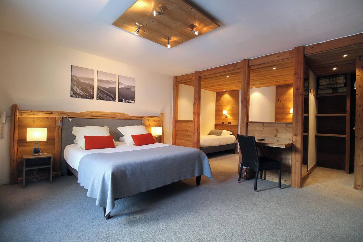 A bedroom with a large bed, a desk, and a chair. There are two bedside tables with lamps. The room has wooden walls and a sloped ceiling with recessed lighting.