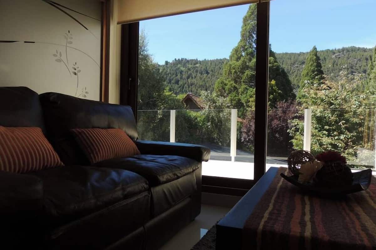 A modern living room with a leather sofa, a coffee table, and a large window offering a view of a forested hillside.