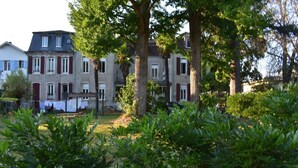 A row of traditional European-style buildings with multiple windows and shutters, surrounded by lush greenery and trees. - Le Trianon de Salies (Salies-de-Bearn)