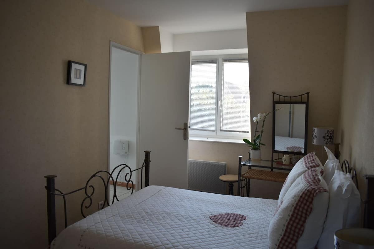 Une chambre à coucher comprenant un lit, une table de chevet, un miroir et une fenêtre avec des stores.