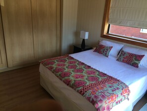 A hotel room with a large bed, a nightstand, a lamp, and a window with blinds. - La Cascada Duplex (San Martín de los Andes)