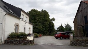 Un camino rural que lleva a un edificio blanco con una entrada de muro de piedra, un coche rojo estacionado al frente y un árbol grande al fondo.
