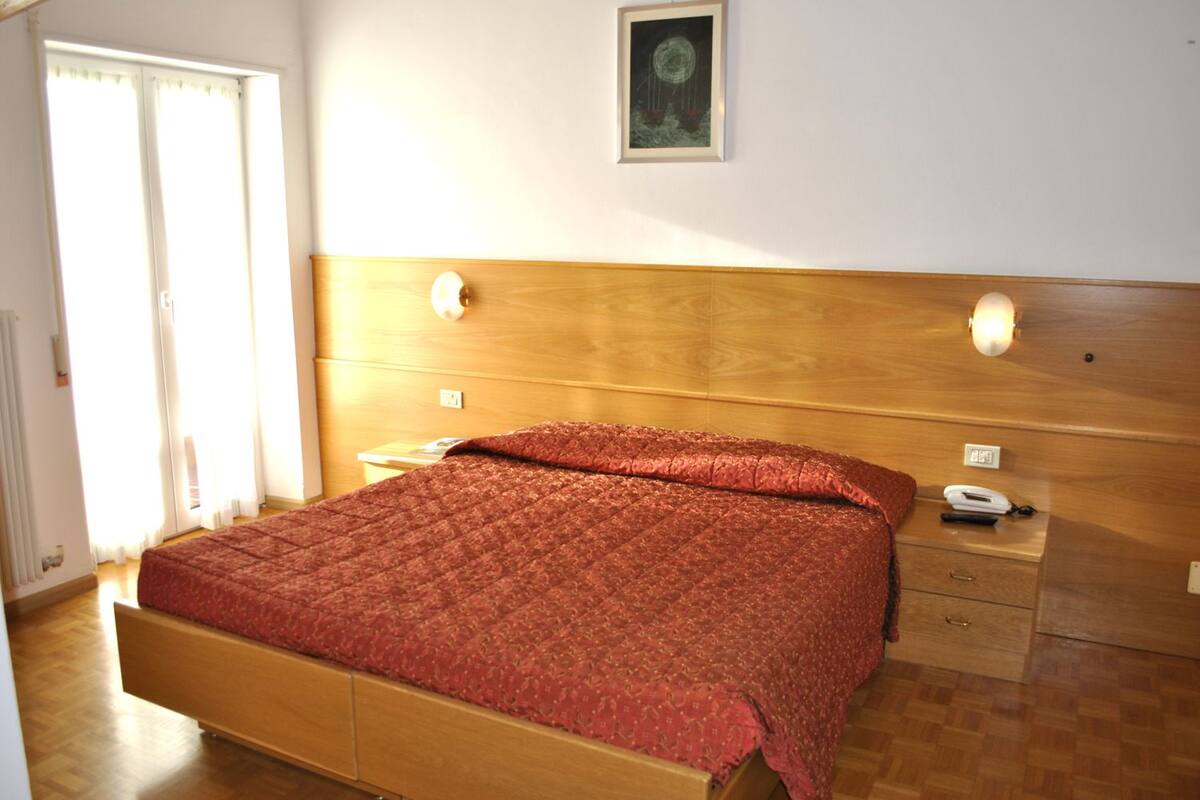 A bedroom with a wooden headboard, a red quilted bedspread, a bedside table with a lamp, and a framed picture on the wall.