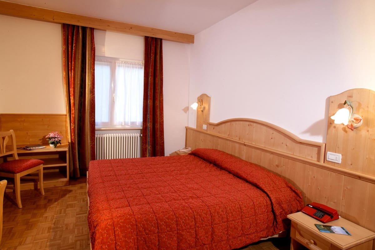 A bedroom with a wooden headboard, a red bedspread, a wooden nightstand with a red telephone, a wooden desk with a chair, and a window with curtains.