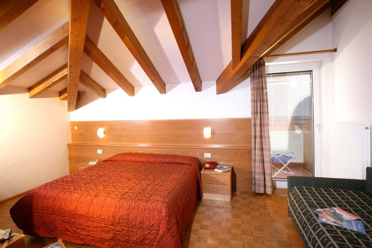 A bedroom with a wooden ceiling, a bed with a red quilt, a bedside table with a book, and a window with blinds.