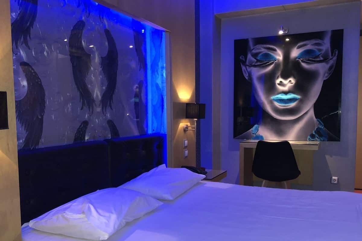 A modern hotel room with a large bed, a wall-mounted TV, and a unique blue-lit artwork.