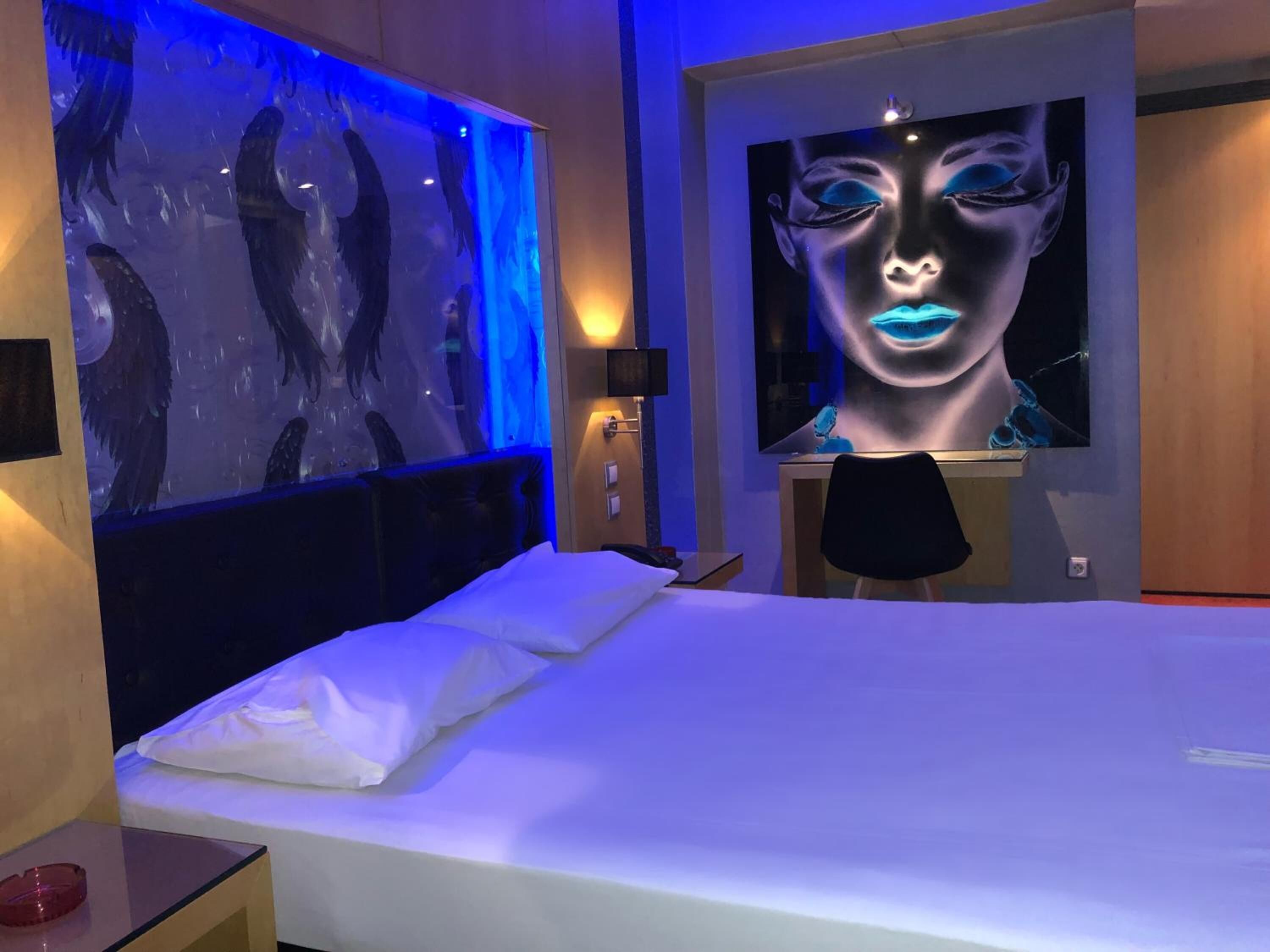 A modern hotel room with a large bed, a wall-mounted TV, and a unique blue-lit artwork on the wall.
