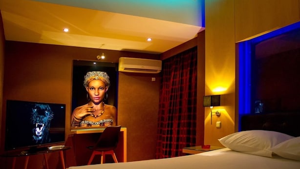 A hotel room with a bed, a desk with a chair, a television, and a wall-mounted air conditioner. - Olympic Hotel – Adults Only (Piraeus)
