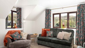 Una sala de estar acogedora con sofá, sillón y una mesita. Amplias ventanas con cortinas, una lámpara y vistas al exterior.