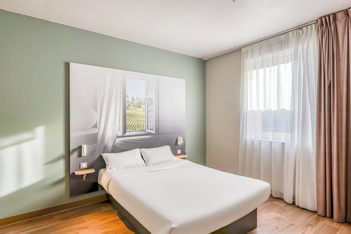 Een net opgemaakt bed in een kamer met een groot raam, een nachtkastje en een kunstwerk aan de muur.