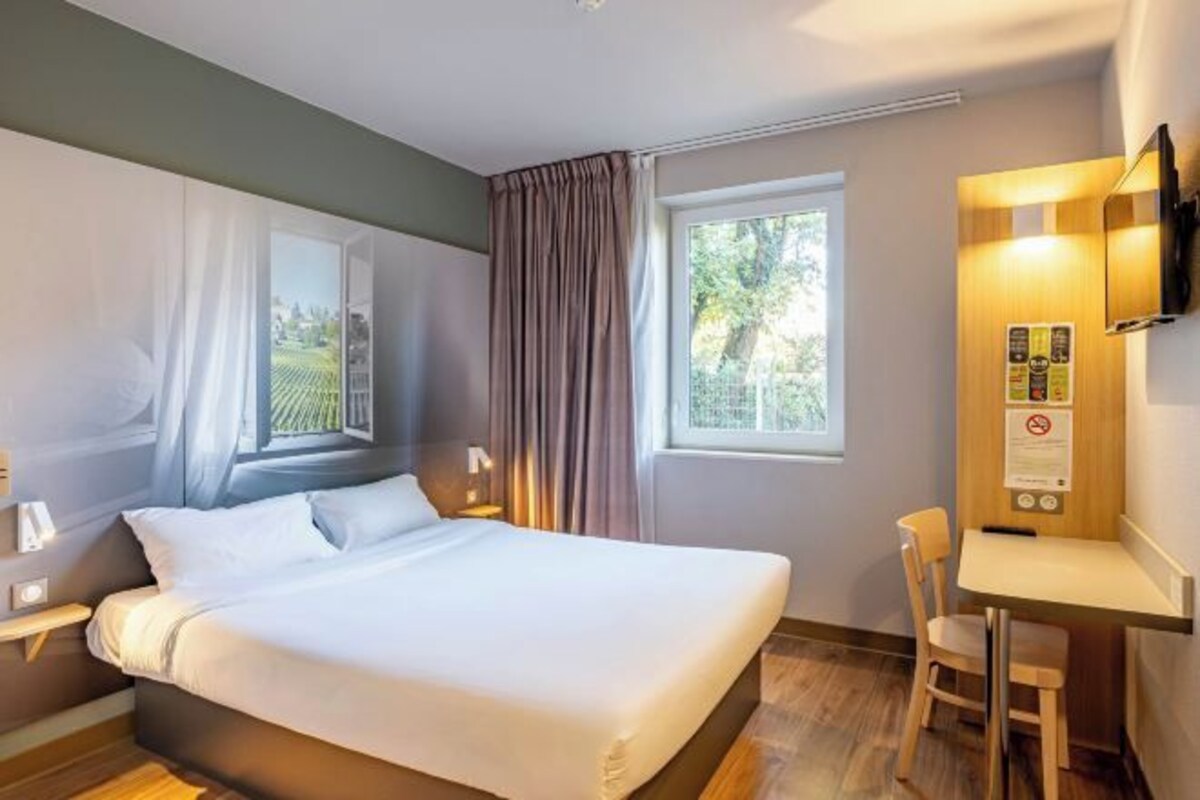 A hotel room with a bed, a desk, a chair, a TV, and a window with curtains.