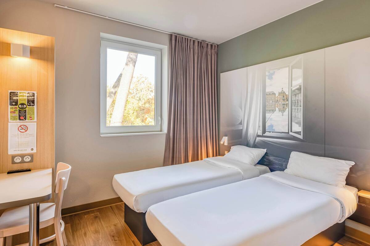 Een hotelkamer met twee eenpersoonsbedden, een bureau met stoel, een raam met gordijnen en uitzicht op een gebouw.