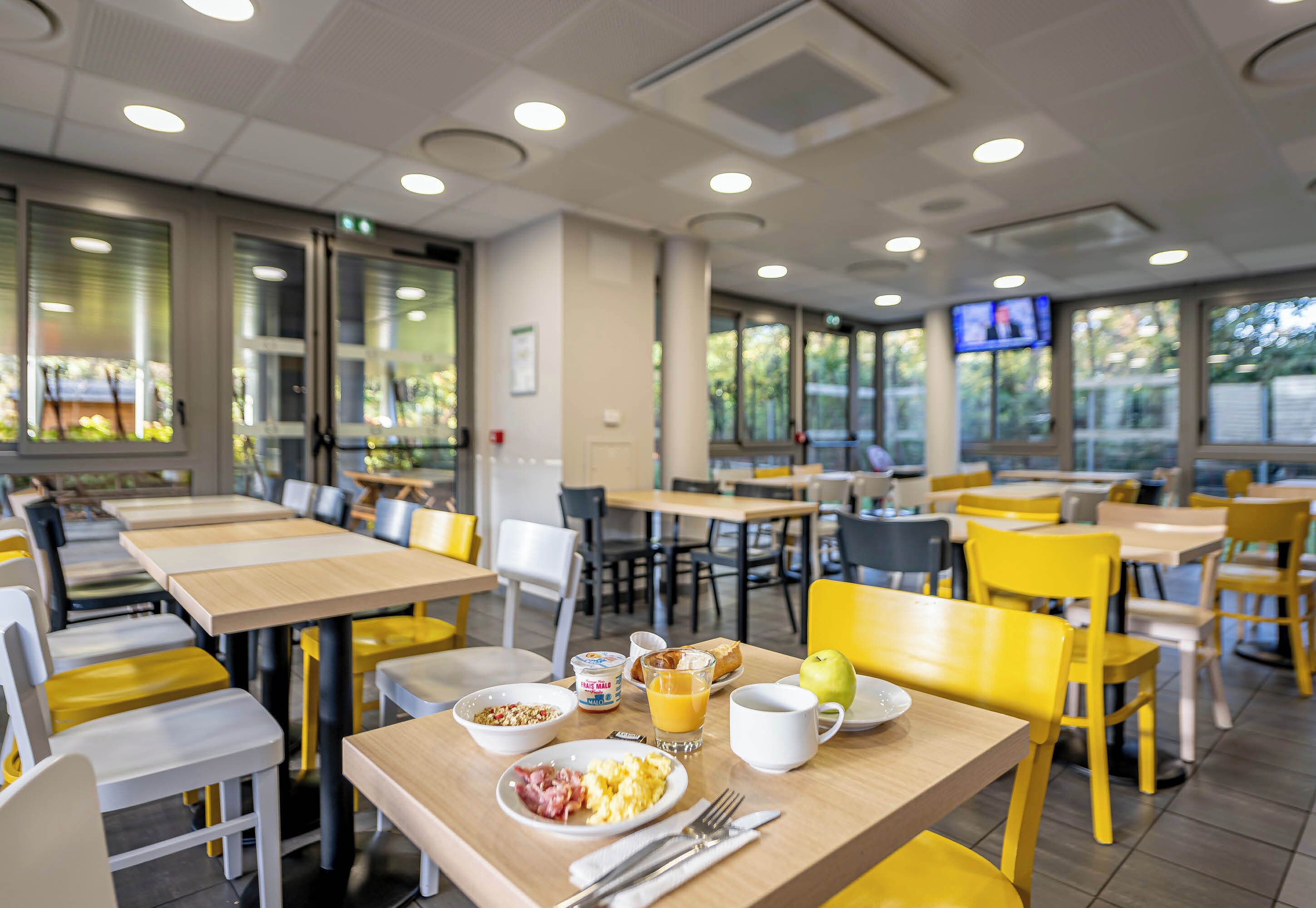 A breakfast setup with a plate of scrambled eggs, bacon, and toast, a glass of orange juice, and a cup on a table in a brightly lit dining area with yellow chairs and tables.